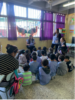 Fotografía de una clase. Docente sentada en una silla y los alumnos en el piso escuchandola.