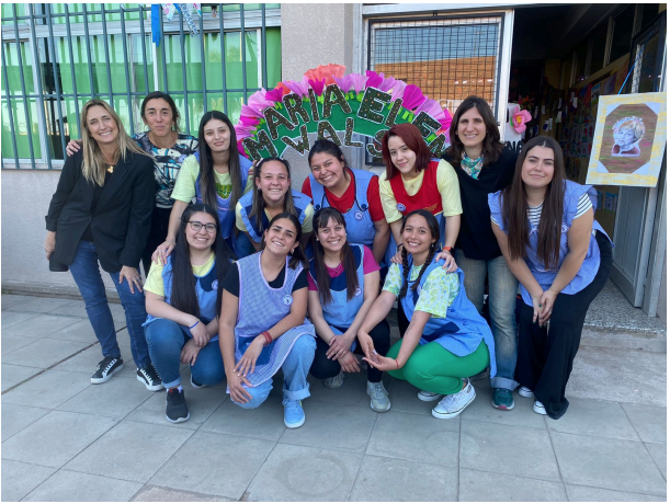Fotografía grupal, de las docentes sonrientes y con una decoración de papel detrás que dice "María Elena Walsh"