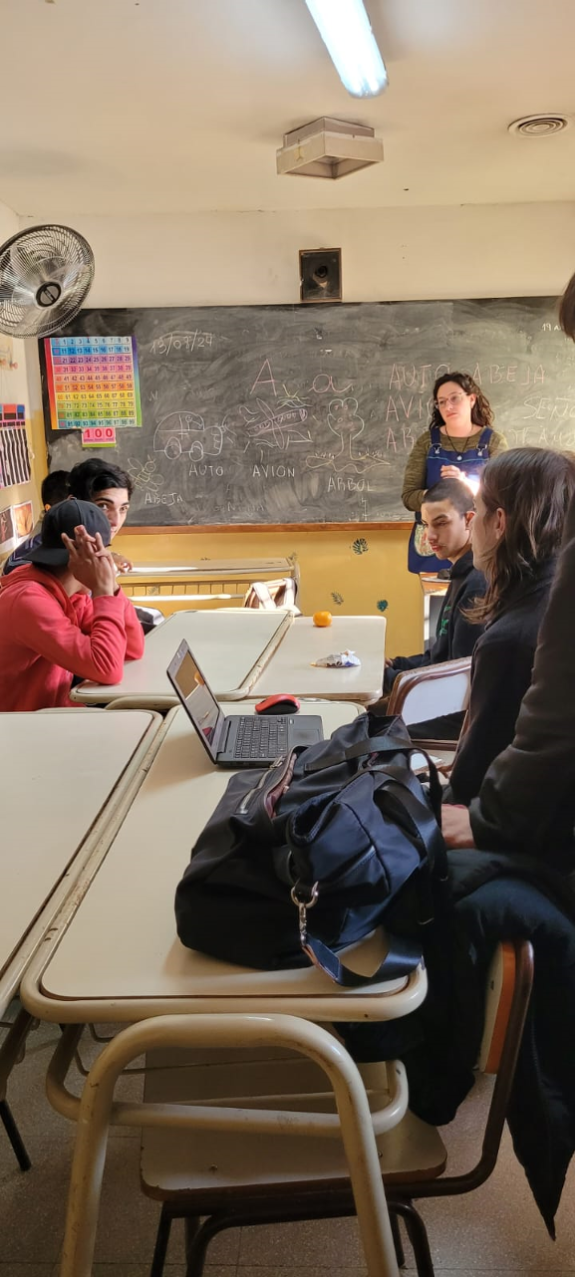 Fotografía en un aula. Docente explicando y estudiantes con una computadora