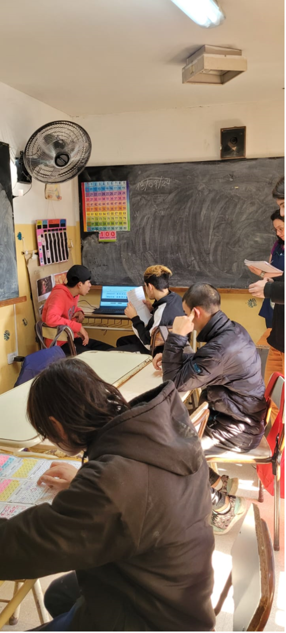Fotografía en un aula. Docente explicando y estudiantes con una computadora