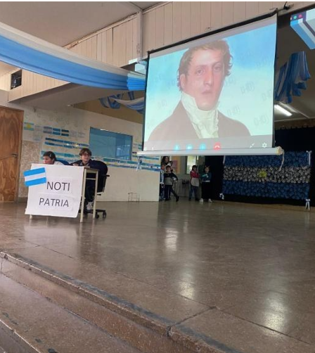 Fotografía del acto. Dos estudiantes exponiendo en una mesa con un texto "Noti Patria", una bandera argentina y detrás la proyección de una imagen de Belgrano