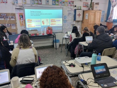 Fotografía de docentes en distintas mesas, trabajando con computadoras. De fondo se ve la proyección de una diapositiva